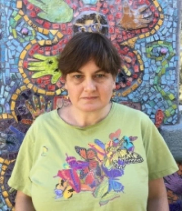 Photo of Anne, an older woman with short brown hair, curtain bangs, and pale skin. She is wearing a light green shirt with the image of a colorful flower on it and standing in front of a mosaic mural of a flower.