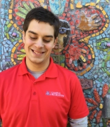 Image of a man with short brown hair and wearing a red polo smiling in front of a colorful mosaic mural.