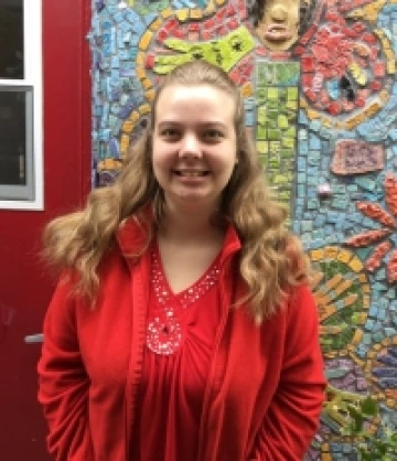 Photo of Carolee, a woman with long, curly ginger hair and wearing a red coat over a red blouse smiling in front of a mosaic mural.