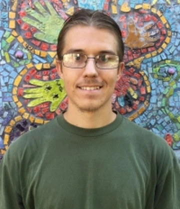 Image of Cody, a man with pulled back brown hair, tan skin, and glasses wearing a green T-shirt and smiling in front of a mosaic mural.