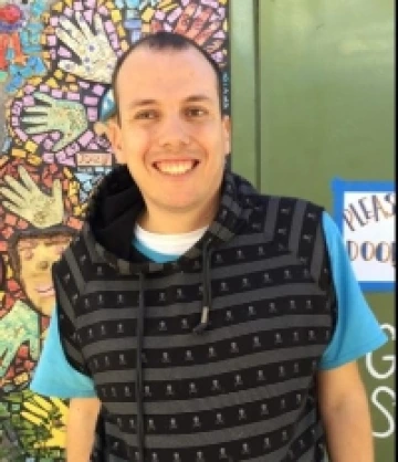Image of Jack, a man with short brown hair waering a black sweater vest over a blue T-shirt, smiling in front of a mosaic mural.