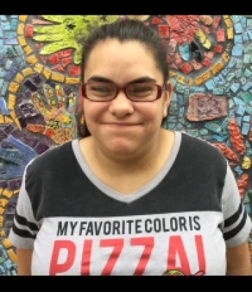 Image of Jennessa, a woman with brown hair in a ponytail, red-framed glasses, an a black and white shirt that says "Pizza" on it, smiling in front of a mosaic mural.