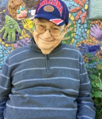 Image of Joe, an elderly man with glasses and wearing a blue and red University of Arizona cap and a black and white wtriped shirt, smiling in front of a mosaic mural.