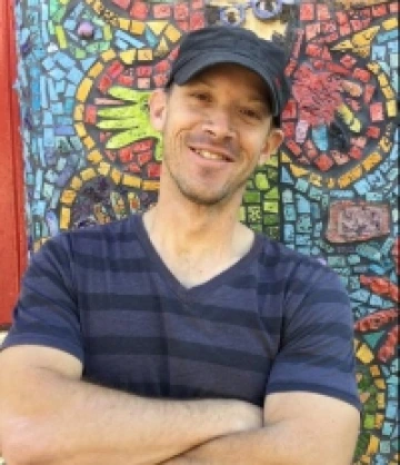 Image of Jon, a man wearing a black baseball cap and wearing a navy and black striped shirt, smiling in front of a mosaic mural.