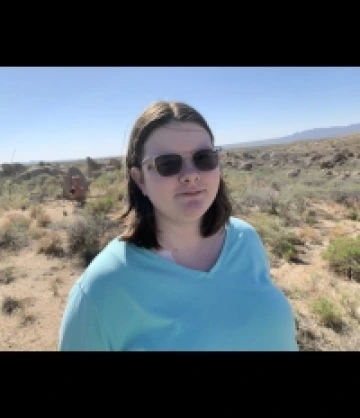 Photo of Madison, a woman with short brown hair and pale skin, wearing square sunglasses and a light blue shirt in a desert brush background.