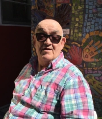 Image of Marco, a bald man wearing sunglasses and a pink and blue plaid shirt, smiling in front of a mosaic mural.