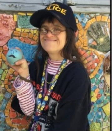 Image of Monica, a woman with long brown hair wearing a black hat and a black long-sleeved shirt, holding a blue ball with a face and a mustache on it, smiling in front of a mosaic mural.