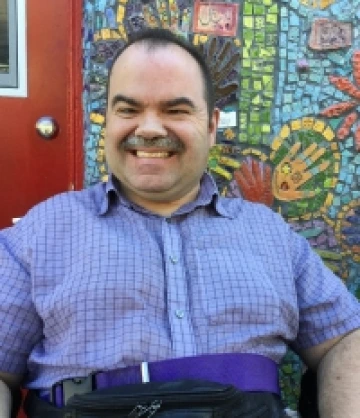 Image of Paul, a man with short brown hair and a mustache, wearing a blue button-down collared shirt, smiling in front of a mosaic mural.