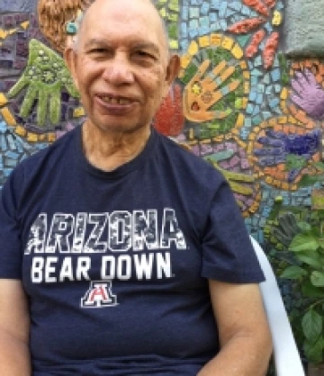 Image of Pete, a bald man with light brown skin and wearing a navy blue shirt that reads "Arizona Bear Down" standing in front of a mosaic mural.