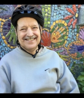 Image of Ron, an elderly man wearing a black bike helment and a grey sweatshirt, smiling in front of a mosaic mural.