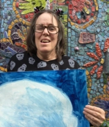 Image of Tina, a woman with shoulder-length brown hair and glasses, wearing a black and white shirt and holding up a blue and white painting while smiling and standing in front of a mosaic mural.