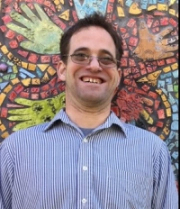 Bradley wearing a blue button up, is wearing glasses and is smiling at the camera