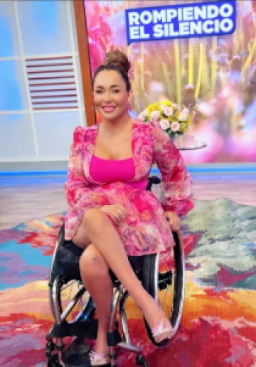 A photo of Tamara Mena, a Hispanic woman with brown and blond highlights with her hair in a bun, wearing a pink dress in a wheelchair.