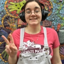 Abby wearing a pink shirt and white overalls with black headphones on and is smiling at the camera