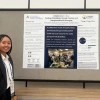 Nadine Chau, an Asian individual with shoulder-length black hair, is standing next to their poster. They are wearing a white collared button-up shirt, a black blazer, and an AUCD lanyard.