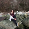 Anahy Fernandez sitting on a large rock smiling. Anahy has long red-brown hair, wearing winter wear, and is holding on to one leg while the other is extended.