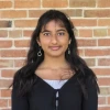 Esha Mahur, wearing a black top, standing in front of a brick wall and smiling