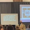 A man speaking at a podium next to a screen showing the IHD Evidence for Success Conference logo.