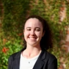  Smiling young woman with curly brown hair pulled back in a ponytail, wearing a black blazer over a white top, standing outdoors in front of a leafy green background 