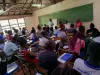 Crowded classroom full of students facing towards a wall with a chalkboard and a man with a red shirt sitting in a wheelchair.