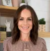 Amber Hansen, a woman with long brown hair, wearing a lavender top with white spots and a gold necklace, standing in front of a shelf and smiling. 