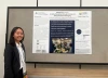 Nadine Chau, an Asian individual with shoulder-length black hair, is standing next to their poster. They are wearing a white collared button-up shirt, a black blazer, and an AUCD lanyard.